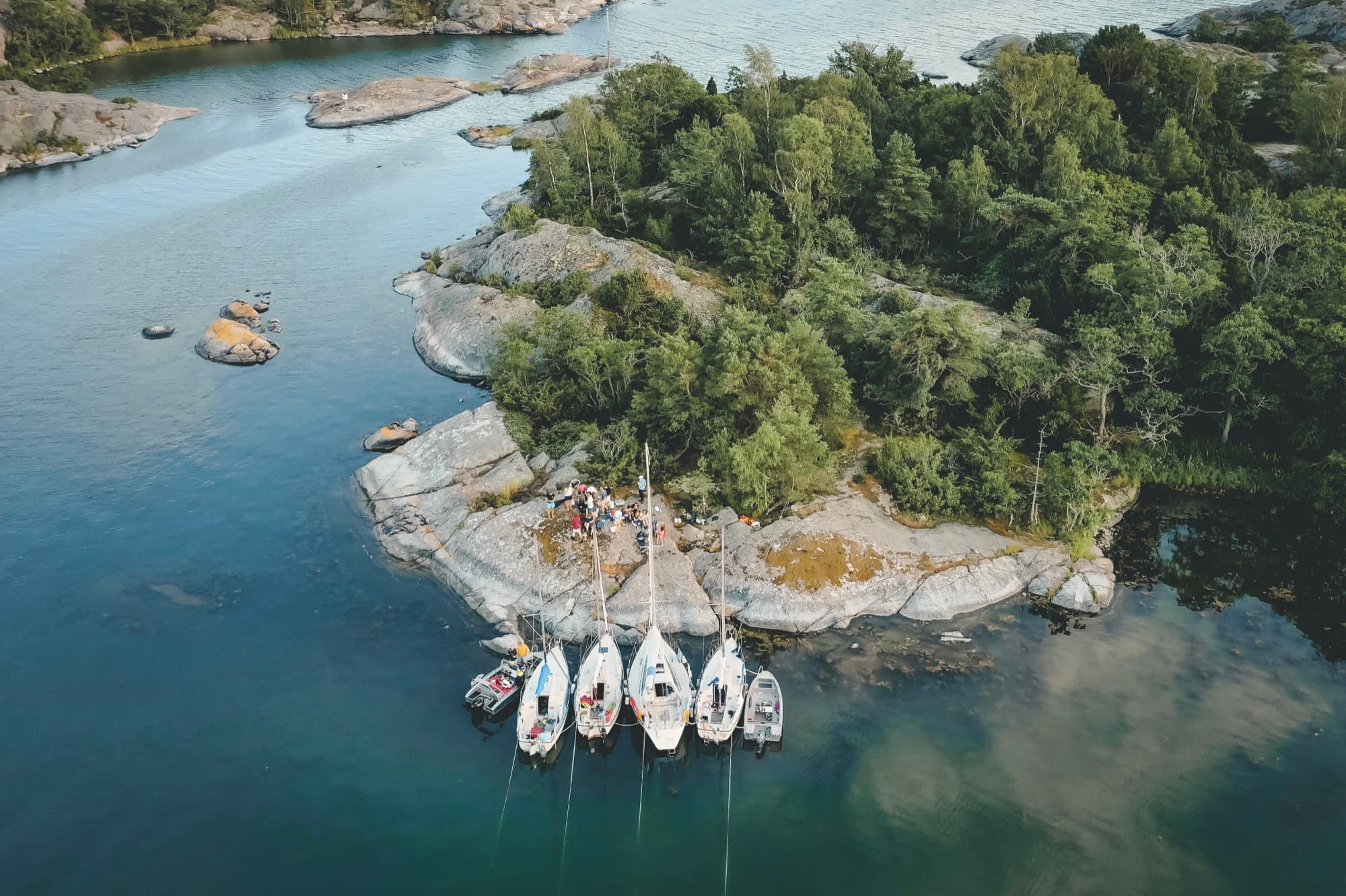 Fem segelbåtar ligger förtöjda vid en klippudde i skärgården, sedda från luften