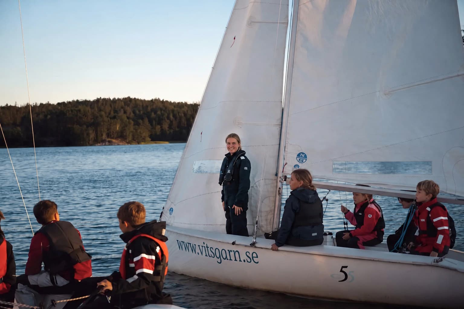 En instruktör står vid seglet på en vit segelbåt medan barn i flytväst sitter ombord